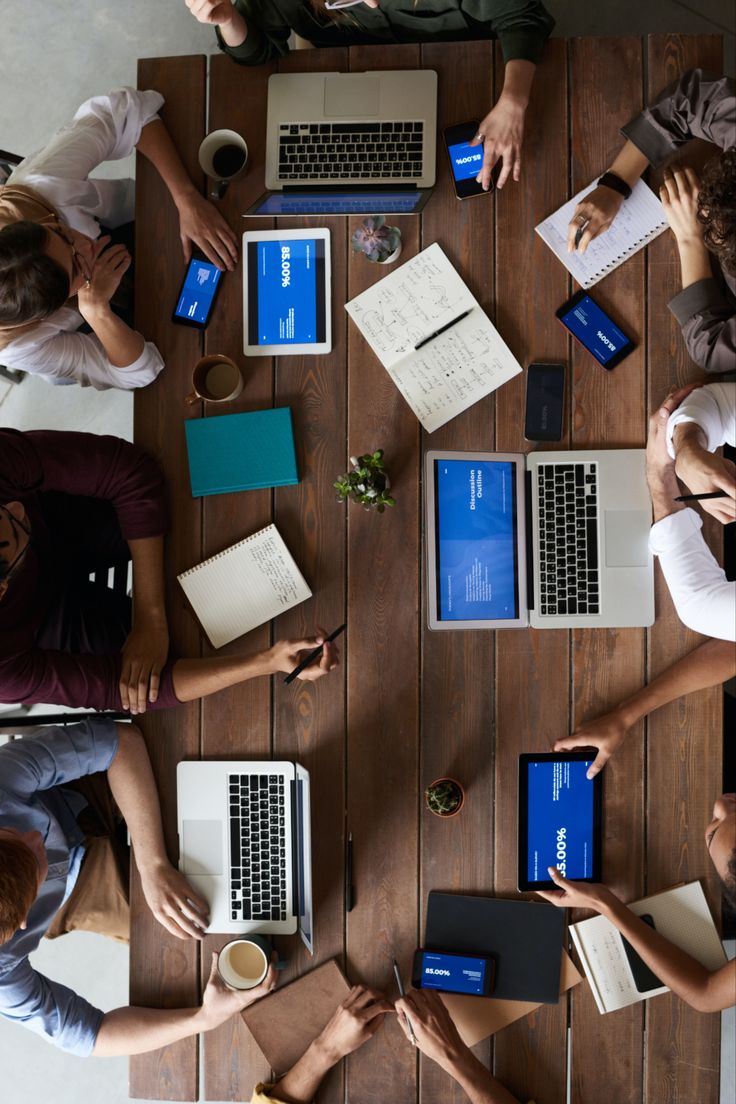 Team collaborating at a wooden desk with laptops, tablets, notebooks, and coffee cups.