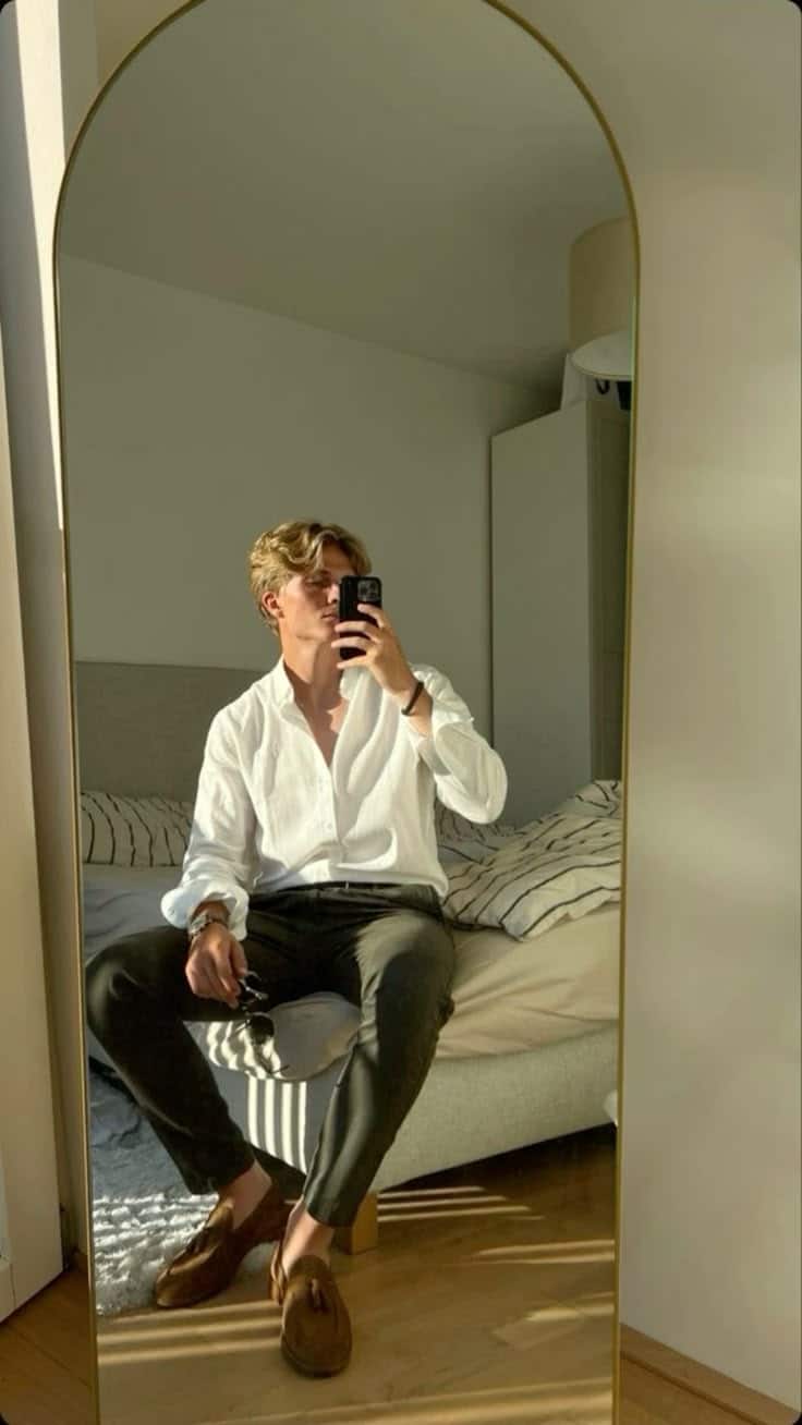 Man in white shirt and sunglasses takes mirror selfie in stylish bedroom.