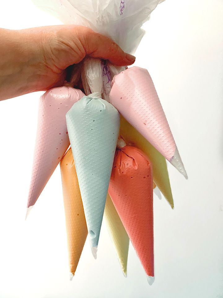 Hand holding six colorful piping bags filled with pastel icing for cake decorating.
