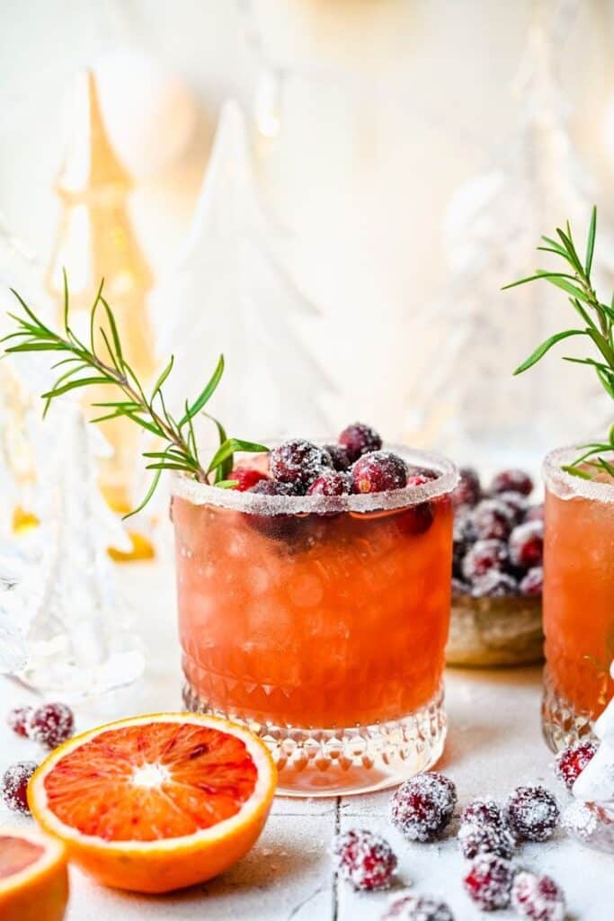 Festive holiday cocktail with cranberries and orange, garnished with rosemary in a glass with sugared rim.