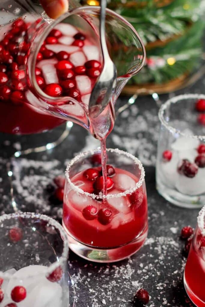 Pouring festive cranberry cocktail into glasses with sugared rims, garnished with fresh cranberries and ice.