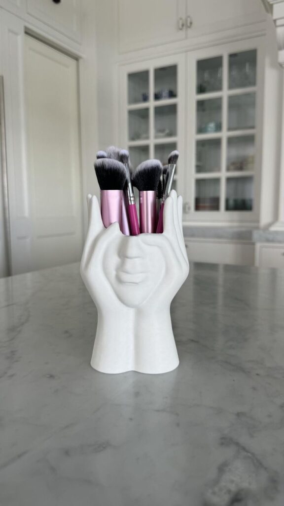 Modern Face Hands Makeup Brush Holder - Unique Decorative Organizer for Vanity or Desk - Stylish Gift for Makeup Lovers White holder with a face design holding pink makeup brushes on a marble countertop. | Sky Rye Design White holder with a face design holding pink makeup brushes on a marble countertop.