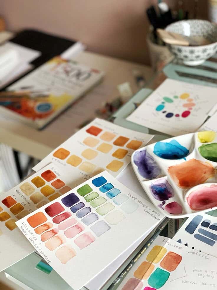 Watercolor palette with color swatches on a cluttered art desk, showing vibrant paint samples and creative tools.
