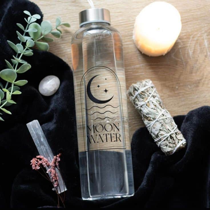 Glass bottle labeled Moon Water with candle and herbs on wooden background.