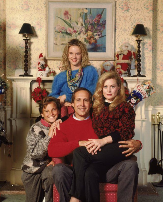 A family poses in festive sweaters with a holiday-themed backdrop, featuring a decorated mantel and Christmas stockings.