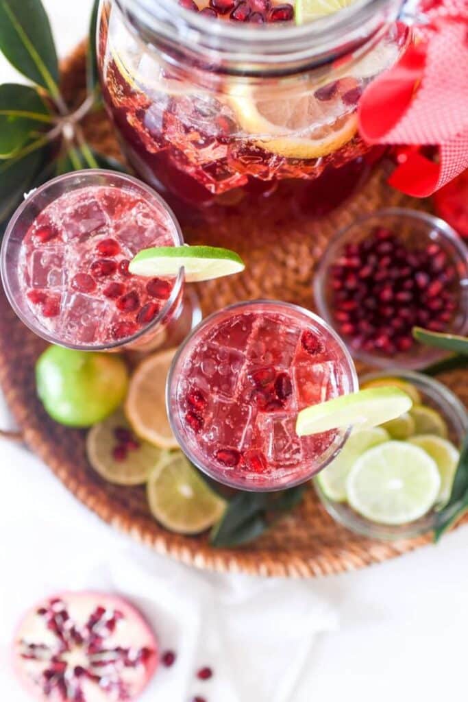 Refreshing pomegranate lime cocktails in glasses with a jug, garnished with fruit slices on a woven tray.