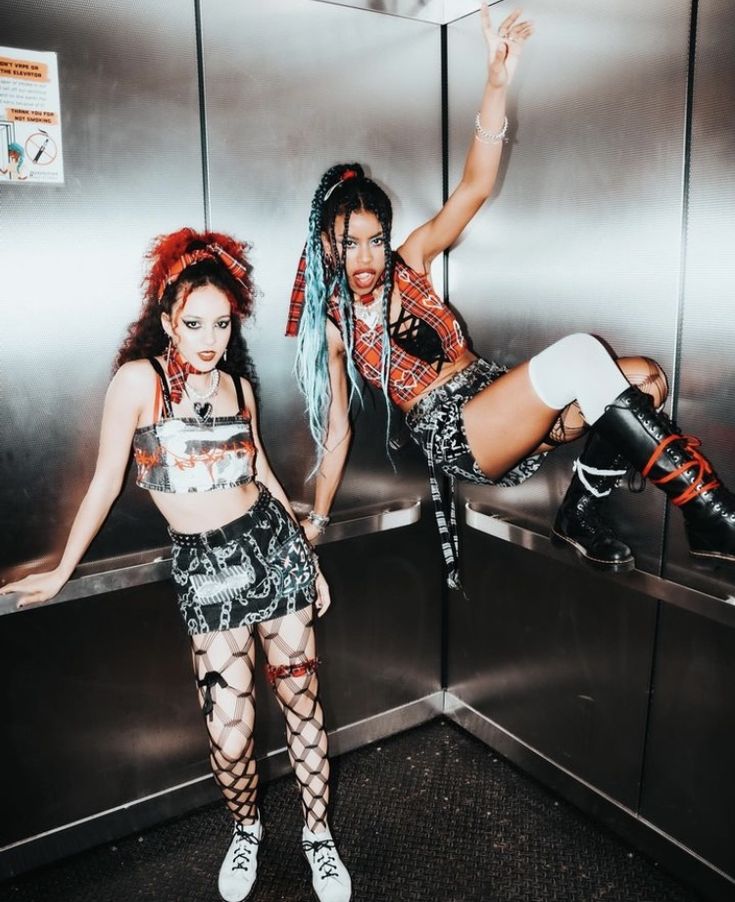Two women in edgy punk outfits pose fiercely in an elevator with metallic walls, showcasing unique style and attitude.