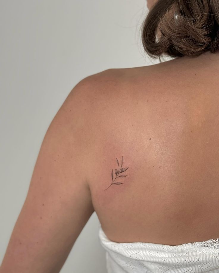 Olive branch back tattoo meaning and design ideas for women Minimalist branch tattoo on woman's upper back wearing white top. | Sky Rye Design Minimalist branch tattoo on woman's upper back wearing white top.