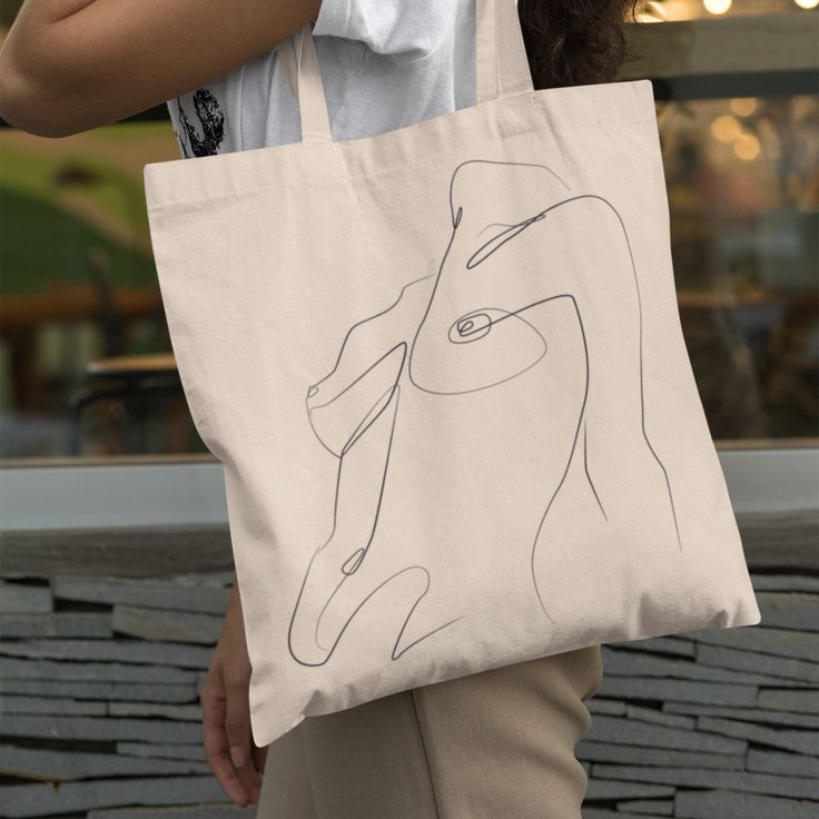 Outfit Bits Bags _ Undressed Female Silhouette Lineart Tote _ Color_ Tan _ Size_ Os Beige tote bag with abstract face line art, carried by person outdoors. Stylish eco-friendly accessory. | Sky Rye Design Beige tote bag with abstract face line art, carried by person outdoors. Stylish eco-friendly accessory.