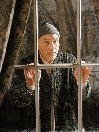 Elderly man in winter hat and robe looks pensively out a frosty window, framed by curtains, in a dimly lit room.