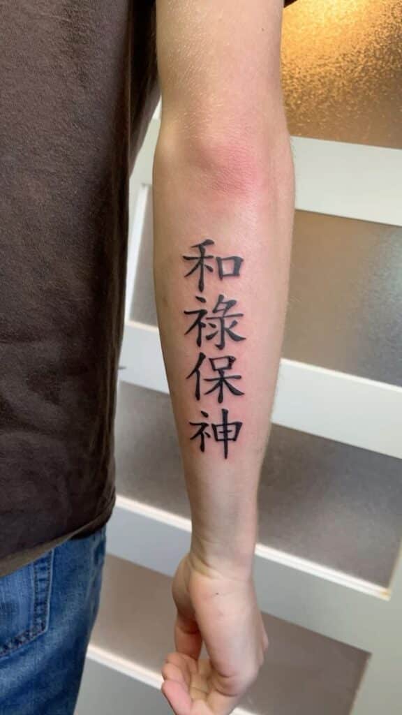 Peace Prosperity Protect God Arm with black Kanji tattoo, featuring four vertical characters, against a neutral background. | Sky Rye Design Arm with black Kanji tattoo, featuring four vertical characters, against a neutral background.