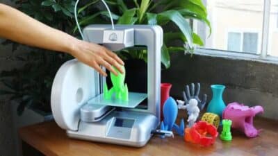 Hand retrieving green 3D printed object from desktop 3D printer, surrounded by colorful 3D printed models.