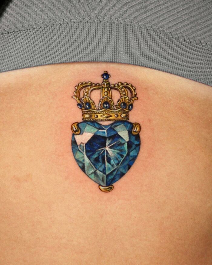 People Go To This Artist For Permanent Jewelry Tattoos Heart-shaped blue gem tattoo with a gold crown design on skin. | Sky Rye Design Heart-shaped blue gem tattoo with a gold crown design on skin.