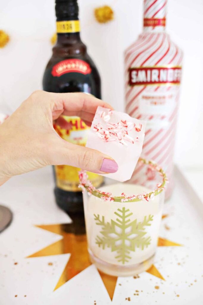 Hand adding peppermint-decorated ice cube to holiday drink with festive glass, and liqueur bottles in the background.