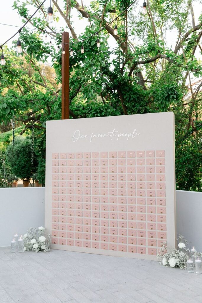 Wedding seating chart display with pink envelopes on a board, adorned with flowers and candles, outdoors.