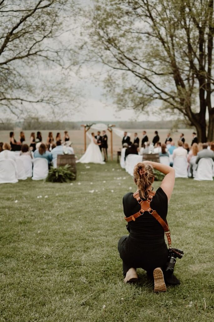 Photographer capturing an outdoor wedding ceremony with guests seated on a grassy lawn under trees.