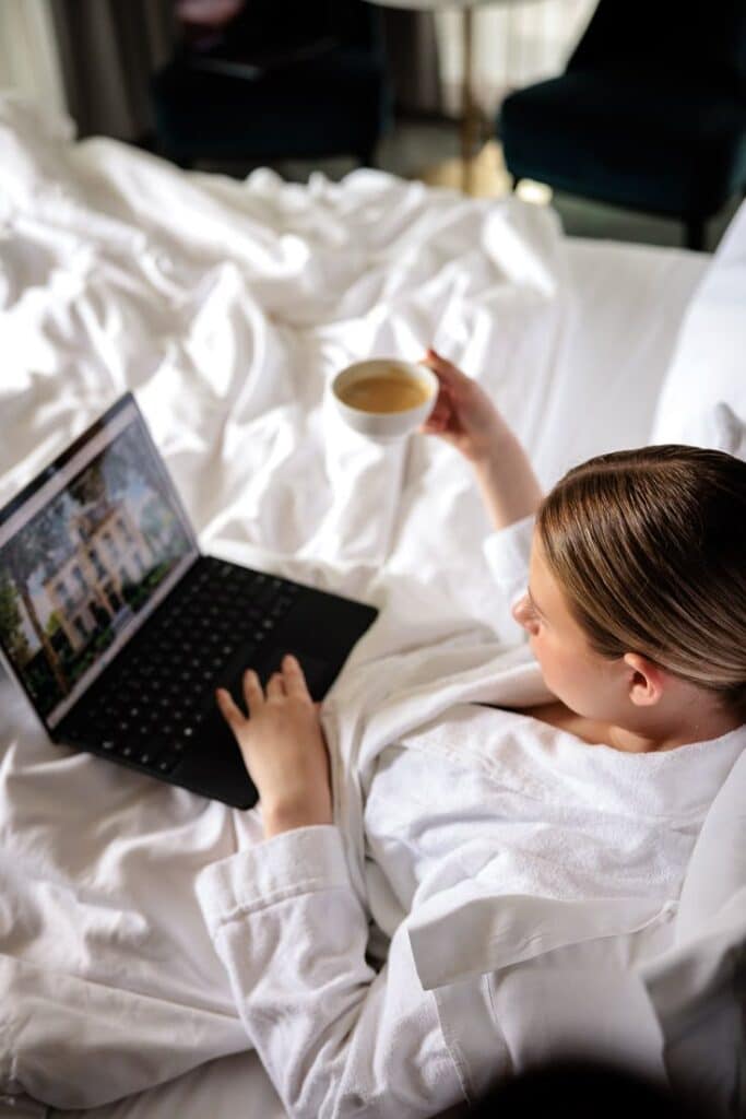 Pillows Grand Boutique Hotel Reylof Ghent - Official website Woman in bed with coffee, using a laptop while relaxing in a white robe. Cozy home office setup. | Sky Rye Design Woman in bed with coffee, using a laptop while relaxing in a white robe. Cozy home office setup.