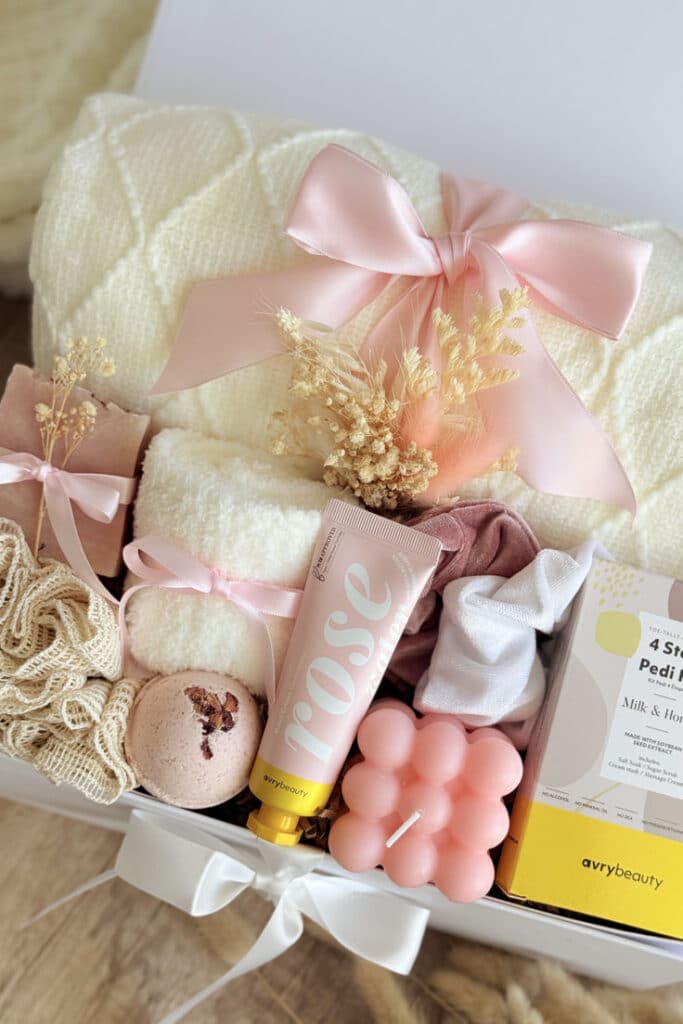 Gift box with cozy items: blanket, candles, skincare, and bath essentials tied with pink ribbons. Perfect for relaxation.