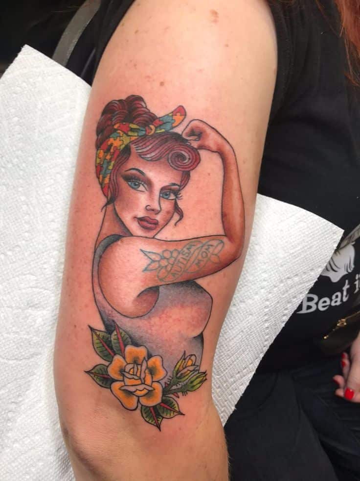 Tattoo of a vintage woman flexing arm with a floral design on upper arm, inspired by Rosie the Riveter.