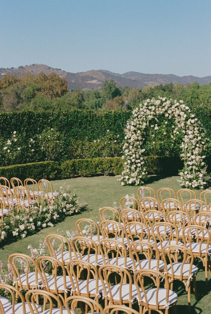 Outdoor wedding setup with floral arch, chairs, and mountain backdrop under clear blue sky. Ideal for romantic ceremonies.