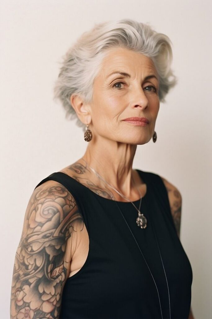 Elderly woman with silver hair and tattooed arms in a black top, wearing earrings, exuding confidence.