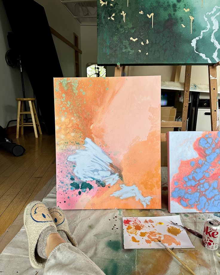 Really enjoying how this new painting is coming Abstract art on canvases in a studio with paint supplies and a person’s feet in smiley slippers visible. | Sky Rye Design Abstract art on canvases in a studio with paint supplies and a person’s feet in smiley slippers visible.