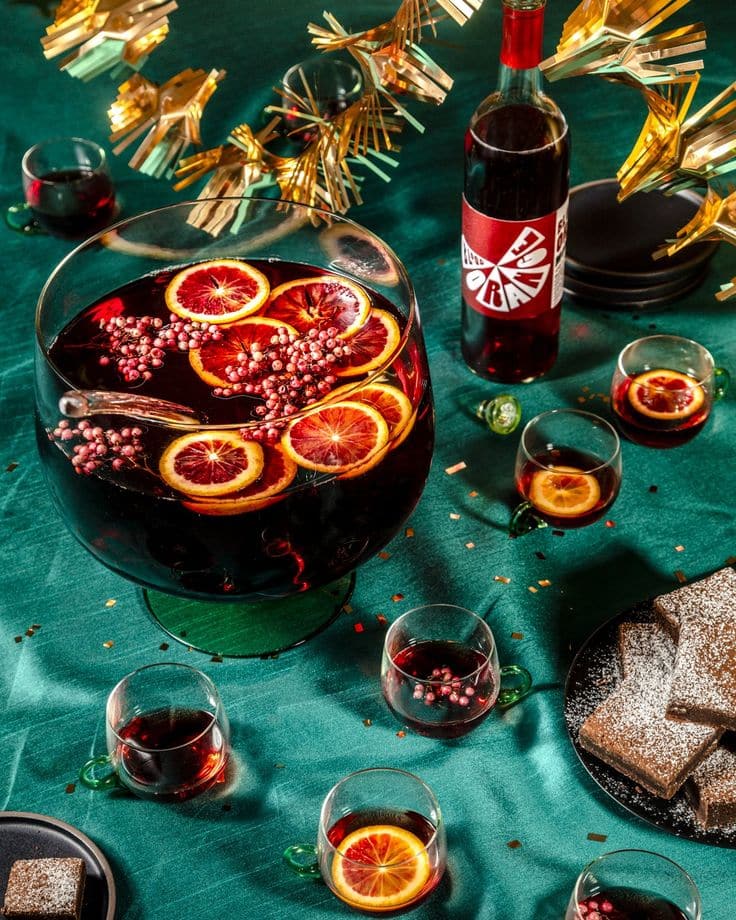 Festive sangria bowl with orange slices and berries on a table, surrounded by glasses and a dessert plate.