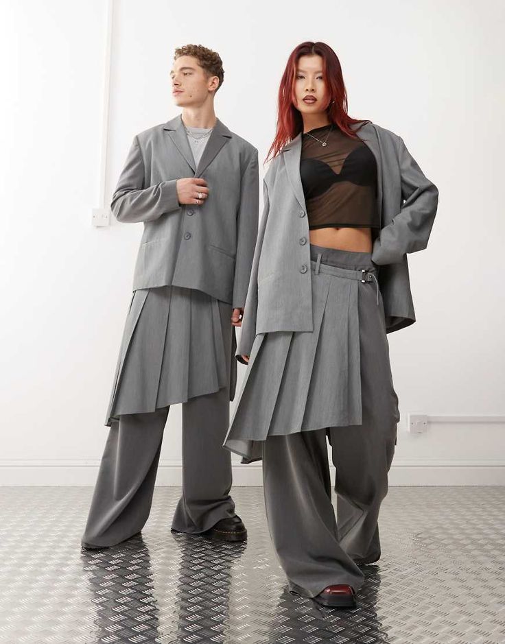 Two models wearing oversized gray suits with layered pleats, standing on a metallic floor in a minimalist setting.