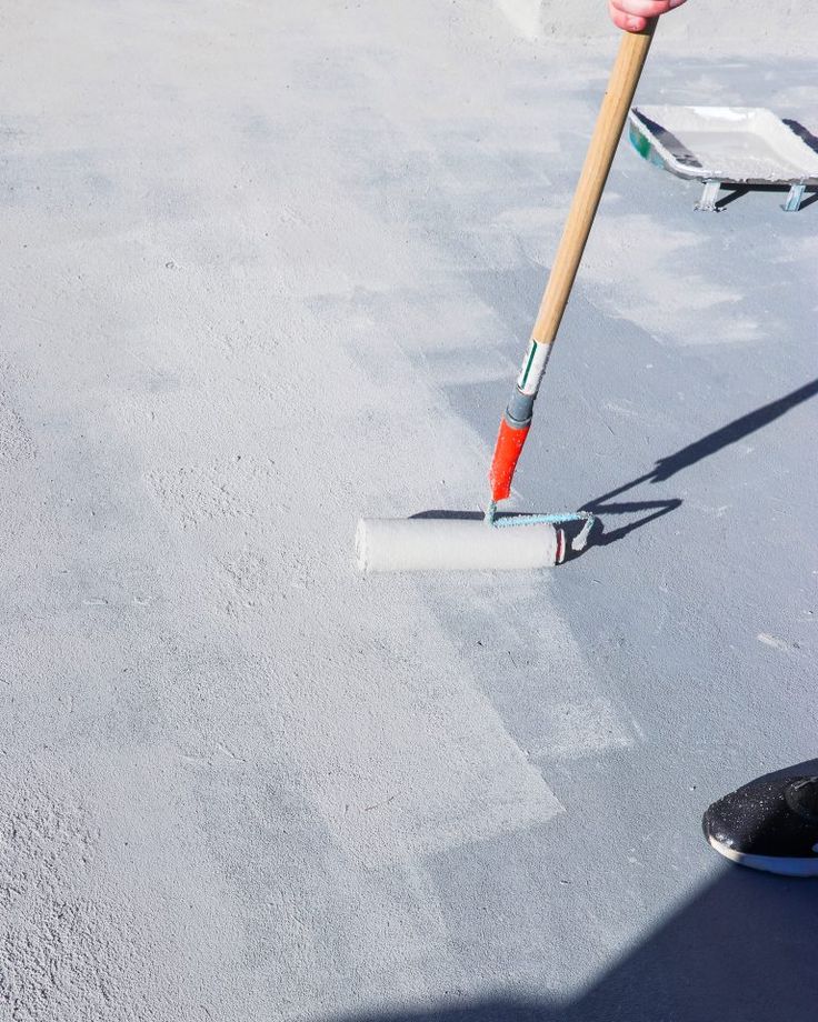 Person applying grey paint on a concrete floor with a roller brush in sunlight.