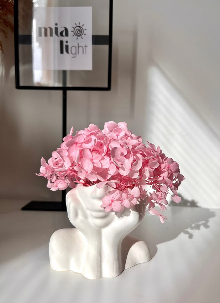 Pink flowers in a unique face-shaped vase, illuminated by sunlight, with a mia light sign in the background.