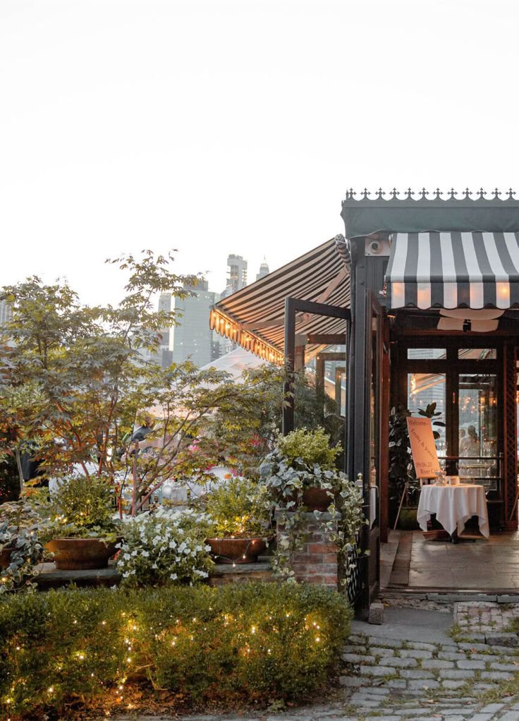 Charming outdoor restaurant with plants and string lights, featuring striped awning and cobblestone path at sunset.