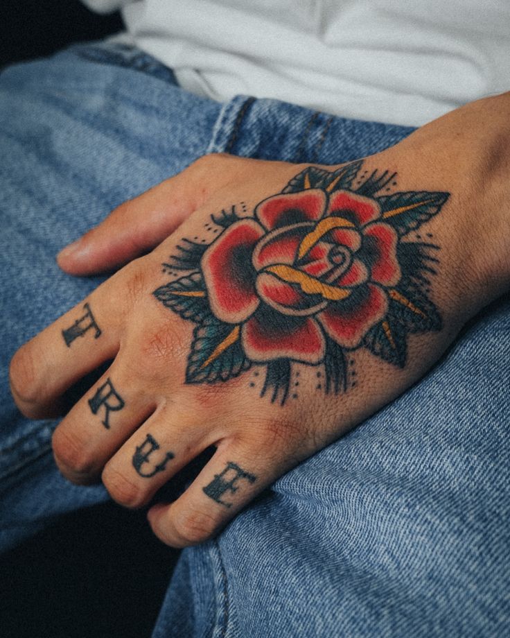 Hand tattoo with a vibrant rose design and TRUE on knuckles, against blue denim.