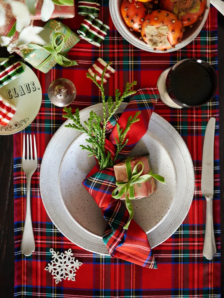 Festive holiday table setting with plaid napkins, greenery, and gift, on a vibrant red and green patterned tablecloth.