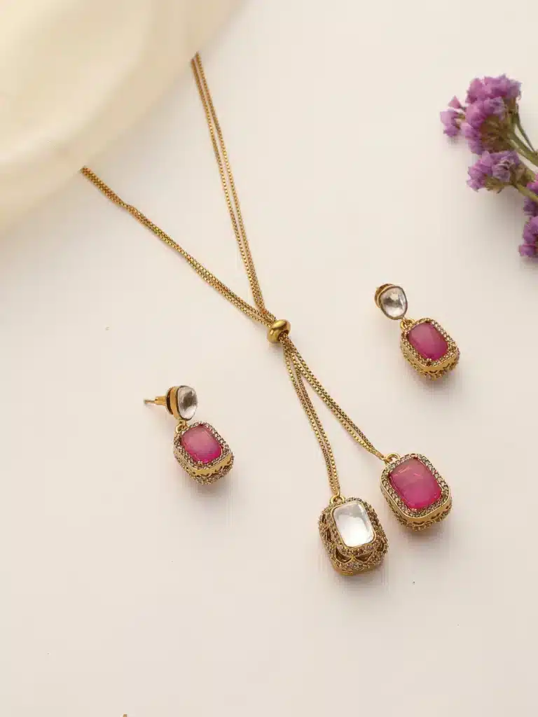 Gold chain with ruby and crystal pendants, matching earrings, on a white background with purple flowers.