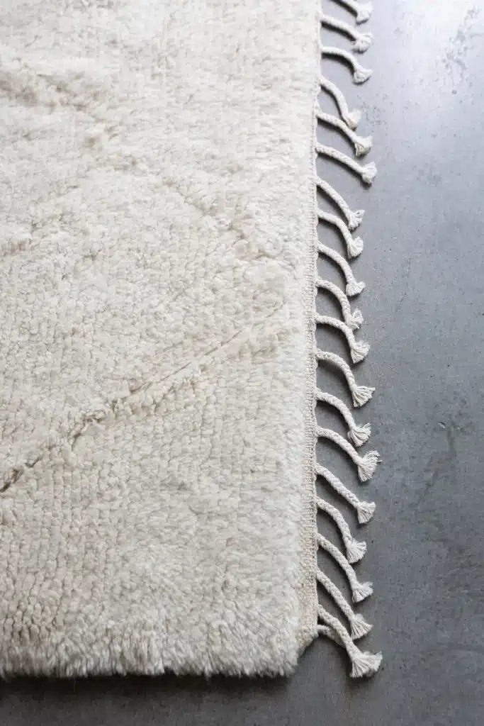 RugsForGood-100_d7a849f7-ab7e-4d37-9d36-b4ab51c361fe_1024x Close-up of a white textured rug with decorative fringes on a smooth gray floor. | Sky Rye Design Close-up of a white textured rug with decorative fringes on a smooth gray floor.