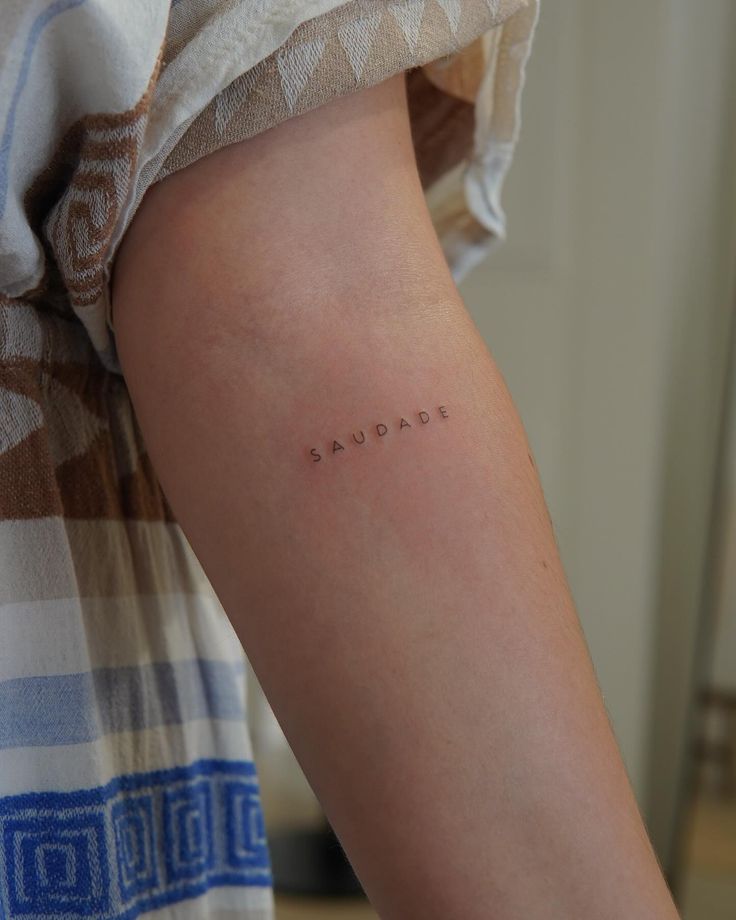 Minimalist Saudade tattoo on forearm, showcasing clean style and precise lettering against a casual outfit background.