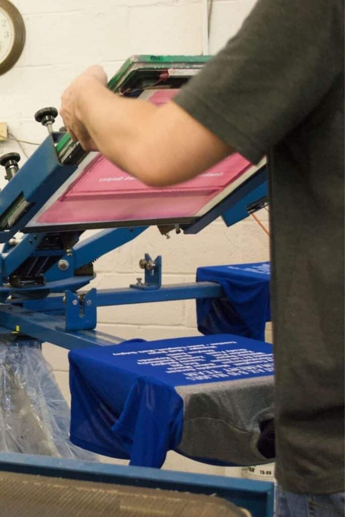 Screen Printing Near Visalia - Convenient Printing Services Person screen printing blue T-shirts in a workshop with manual press machine. | Sky Rye Design Person screen printing blue T-shirts in a workshop with manual press machine.