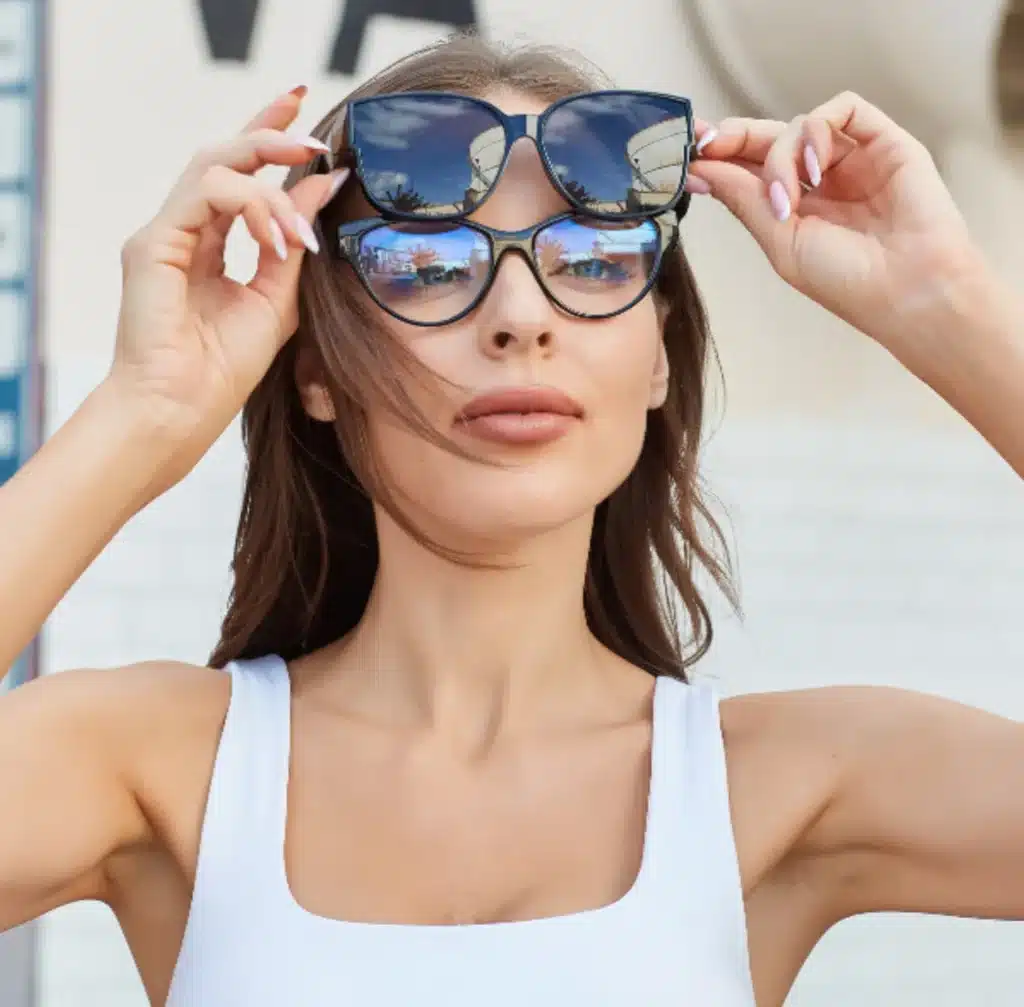 Screenshot_2025-02-13_at_160526 Woman wearing two pairs of sunglasses, holding the top pair. Fashionable eyewear in sunny outdoor setting. | Sky Rye Design Woman wearing two pairs of sunglasses, holding the top pair. Fashionable eyewear in sunny outdoor setting.