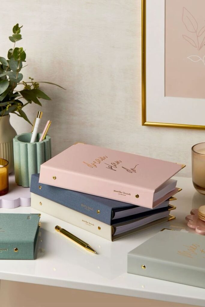 Elegant pastel notebooks and pens on desk, framed art and greenery in the background.