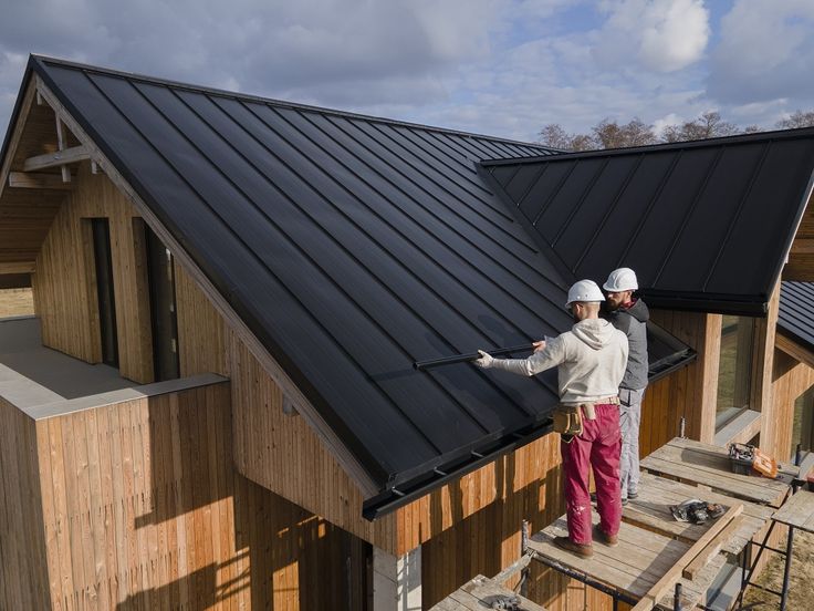 Seamless Rain Gutter Installation Services Construction workers installing a sleek black metal roof on a wooden house under a cloudy sky. | Sky Rye Design Construction workers installing a sleek black metal roof on a wooden house under a cloudy sky.