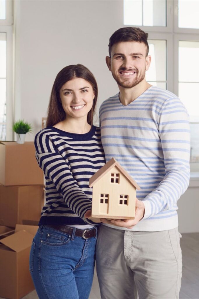 Should you invest in real estate_ Happy couple holding a small wooden house model, symbolizing new homeownership and moving in. | Sky Rye Design Happy couple holding a small wooden house model, symbolizing new homeownership and moving in.