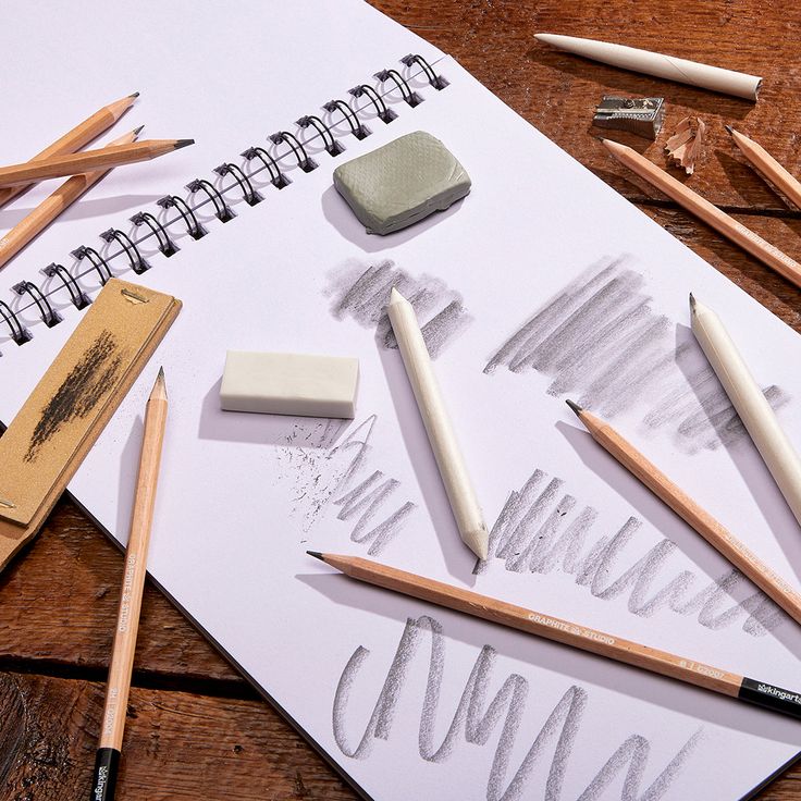 Art supplies on sketchbook: pencils, erasers, blending tools on a wooden table, showcasing sketching techniques.