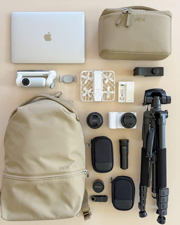 Flat lay of travel essentials: backpack, laptop, camera gear, drone, tripod, and accessories on a beige background.