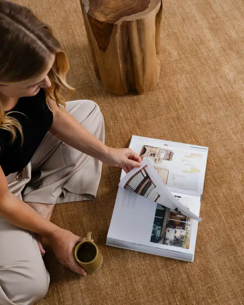 | Sky Rye Design Woman reading a design magazine with tea on a carpet, next to a wooden stool. Cozy and relaxing atmosphere.