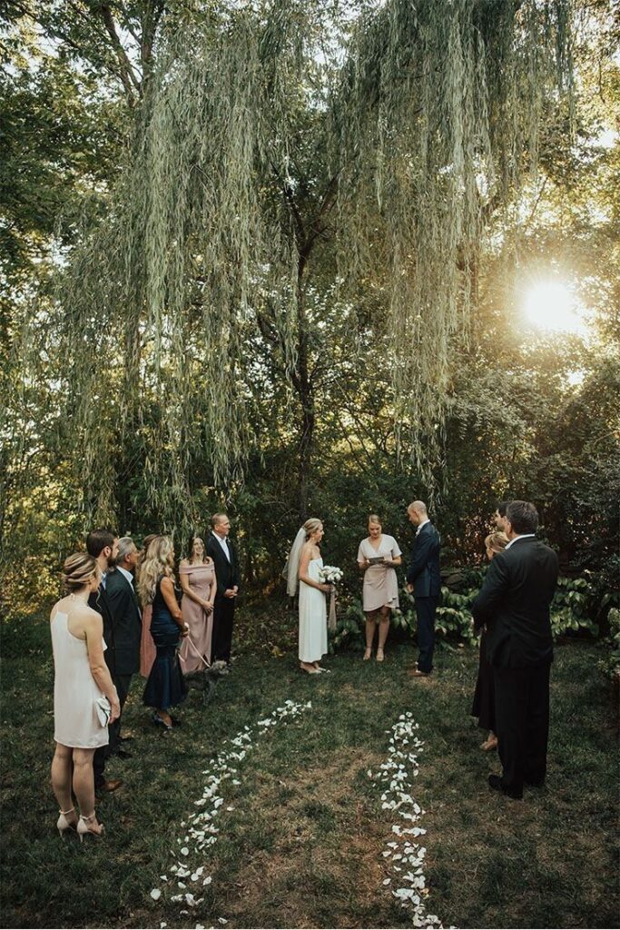 Outdoor wedding ceremony under trees with guests and couple exchanging vows. Romantic garden setting at sunset.