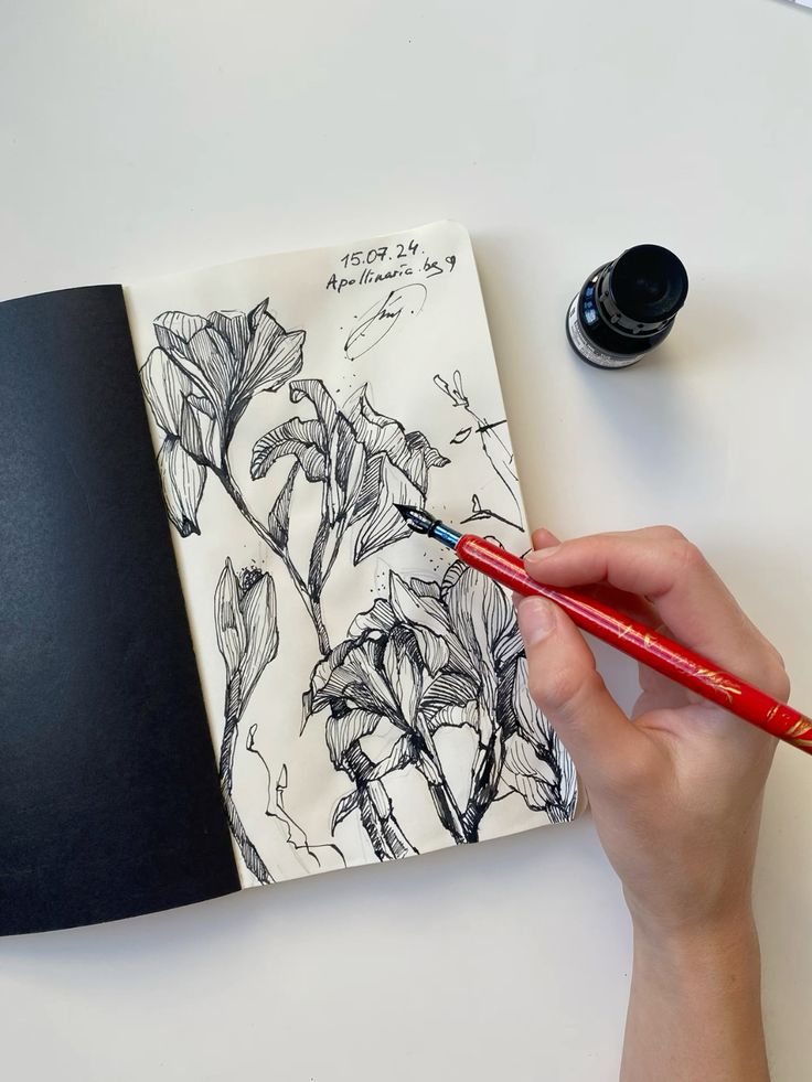 Hand sketching flowers in a notebook with a red pen on white paper, ink bottle nearby.