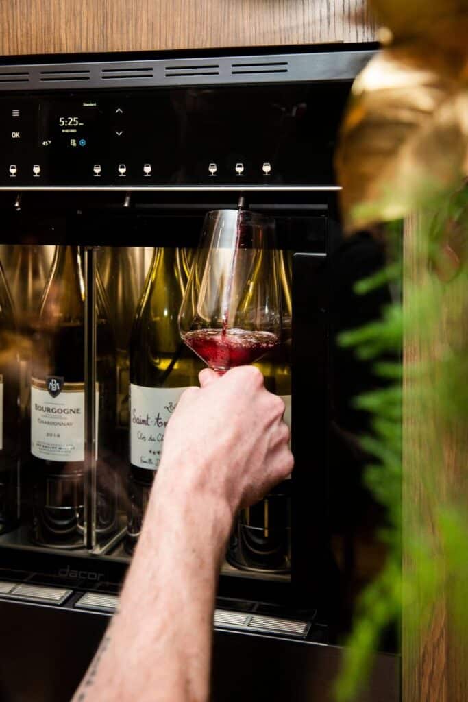 Store Wine Safely With Dacors 24_ Built-in Wine Dispenser Hand pouring red wine from dispenser; wine bottles visible in background. | Sky Rye Design Hand pouring red wine from dispenser; wine bottles visible in background.