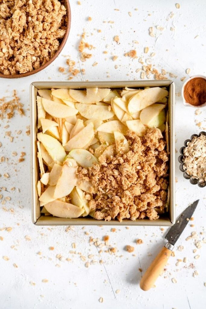 Super Easy Apple Crumble Apple Crisp Square pan with sliced apples and crumble topping, surrounded by oats, cinnamon, and a knife on a white surface. | Sky Rye Design Square pan with sliced apples and crumble topping, surrounded by oats, cinnamon, and a knife on a white surface.