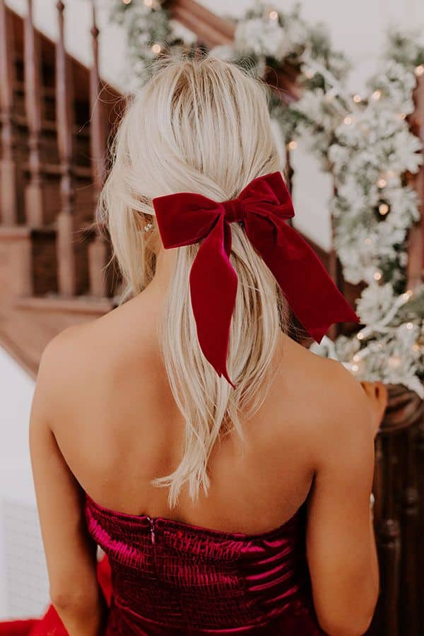 THE BAILEE VELVET BOW HAIR CLIP IN WINE Blonde woman in red dress with velvet bow, standing by decorated staircase during holiday season. | Sky Rye Design Blonde woman in red dress with velvet bow, standing by decorated staircase during holiday season.