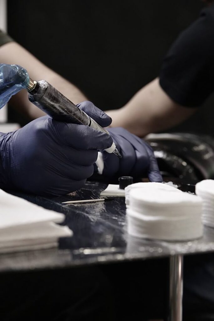 Tattoo 1 Tattoo artist with gloves tattooing, surrounded by ink and cotton pads on a table. | Sky Rye Design Tattoo artist with gloves tattooing, surrounded by ink and cotton pads on a table.